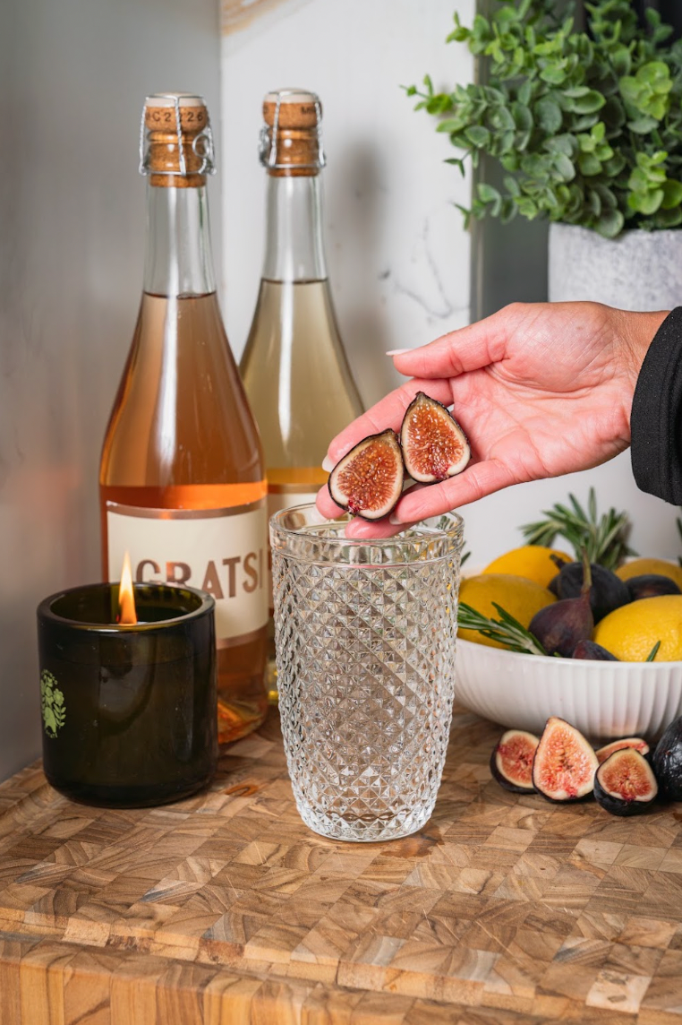 A hand holds two fig halves over an empty textured glass, with wine bottles, a lit candle, and a bowl of lemons and figs nearby on a wooden surface.