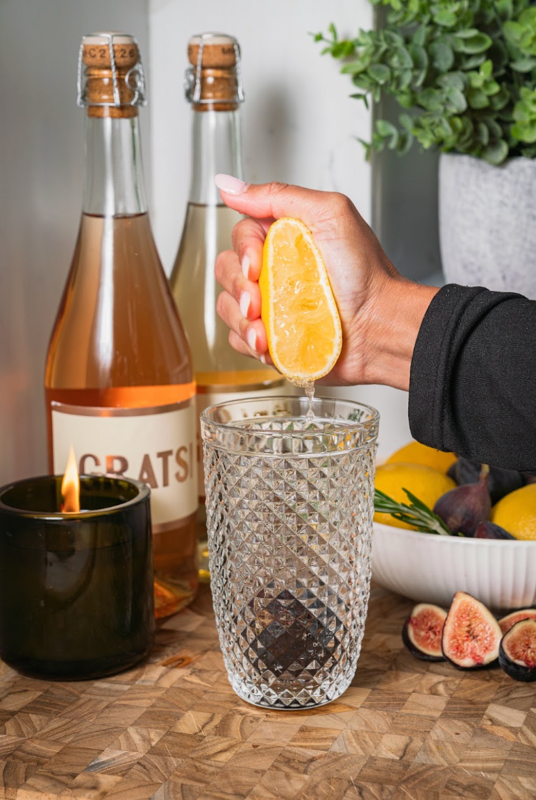 A hand squeezes a lemon wedge into a textured glass, with wine bottles, a lit candle, and fresh figs nearby.