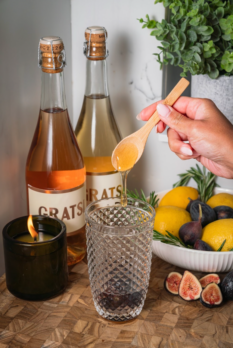 A hand drizzles honey from a wooden spoon into a textured glass, with two bottles of Gratsi wine, a bowl of lemons and figs, and a lit candle nearby.