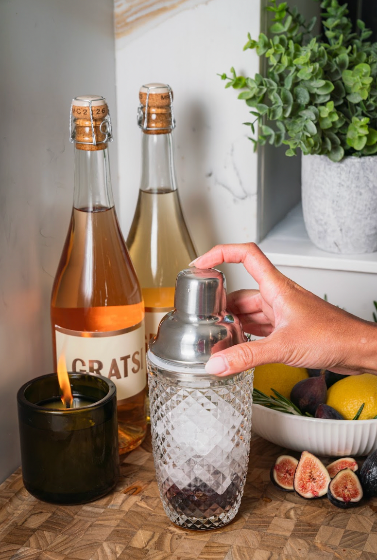 A hand places a metal cap onto a glass cocktail shaker filled with ice, next to wine bottles, a lit candle, and a bowl of figs and lemons.