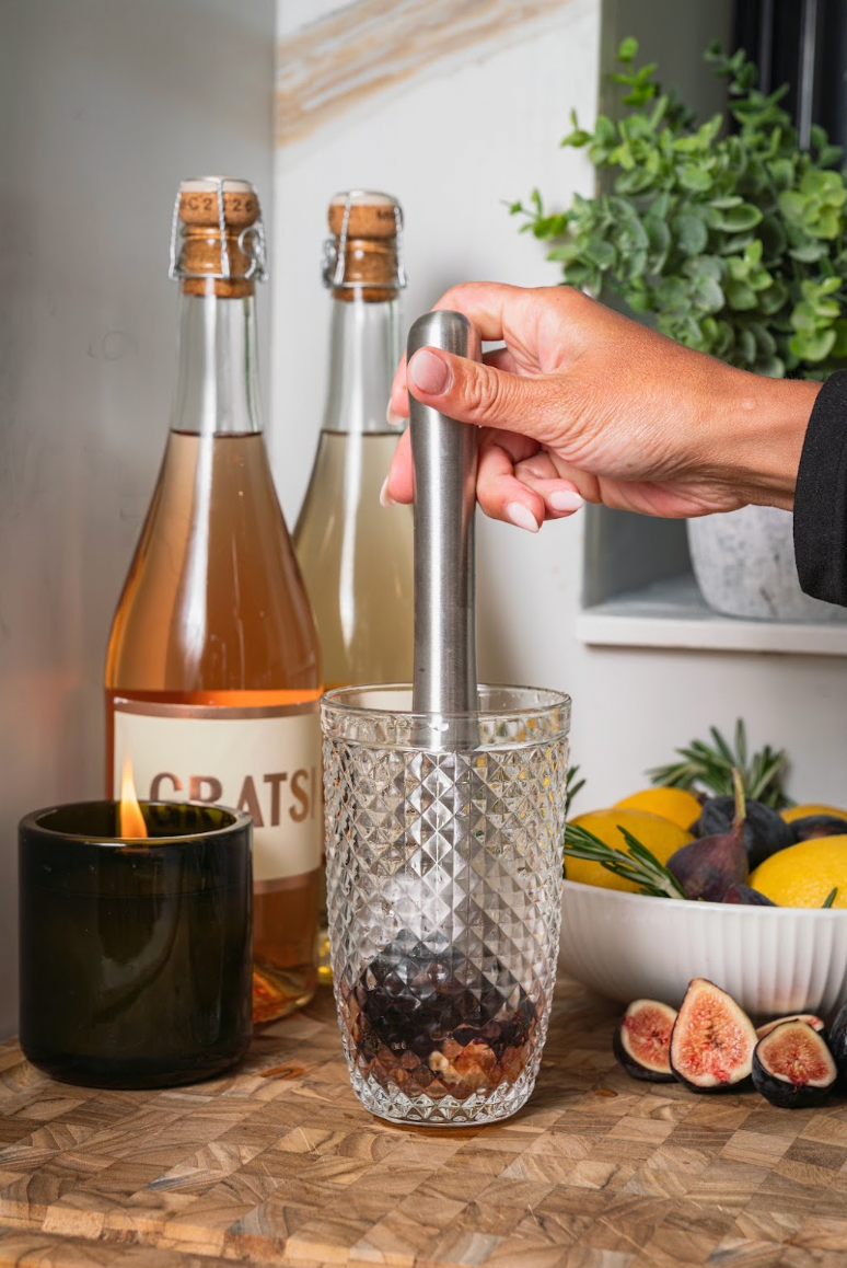 A hand uses a metal muddler to crush figs inside a patterned glass, with two wine bottles, a lit candle, and a bowl of fresh figs nearby.