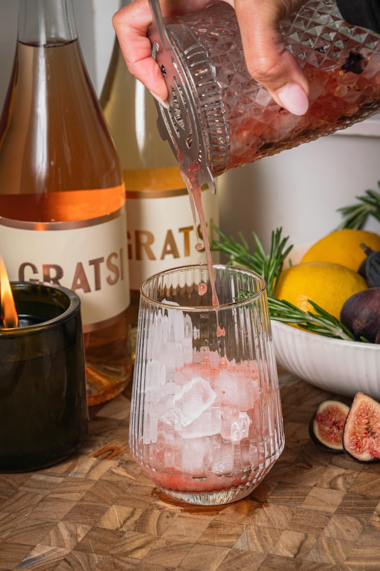 A hand pours a pink cocktail from a glass shaker into a ribbed glass filled with ice, set on a wooden surface with bottles of rosé, lemons, figs, and a lit candle.