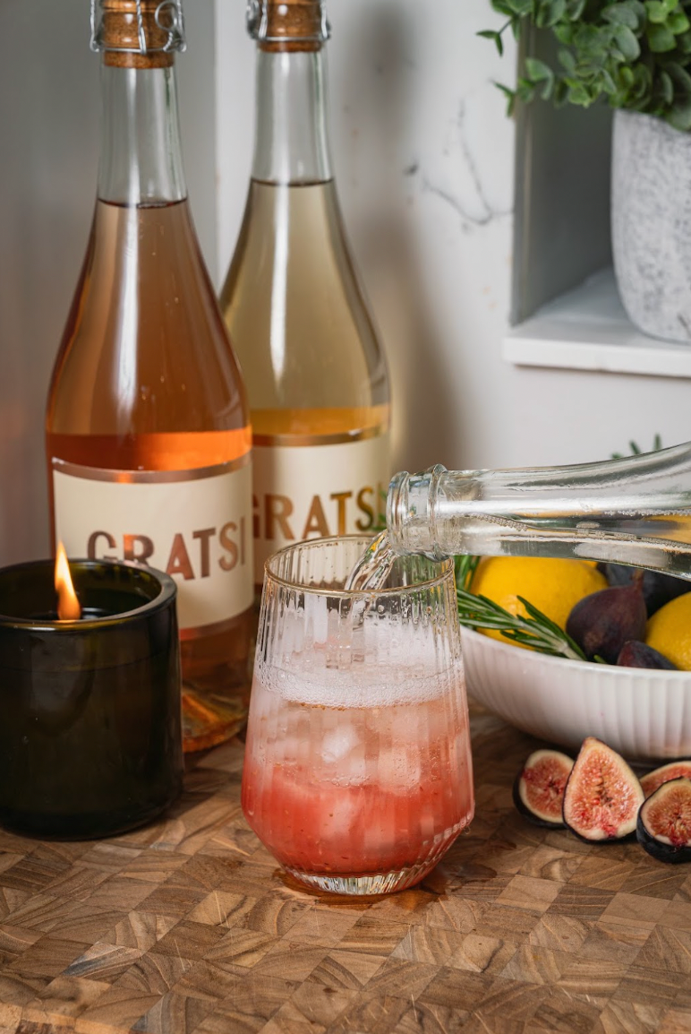 A clear liquid is poured from a bottle into a glass containing a pink drink with ice, with two wine bottles, a lit candle, and fresh figs nearby on a wooden surface.