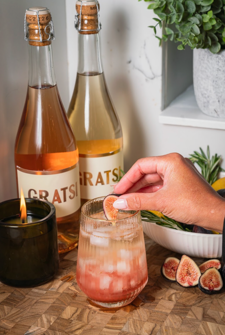 A hand garnishes a glass of iced cocktail with a fresh fig slice, positioned in front of two Gratsi wine bottles, a lit candle, and a bowl of fruit.