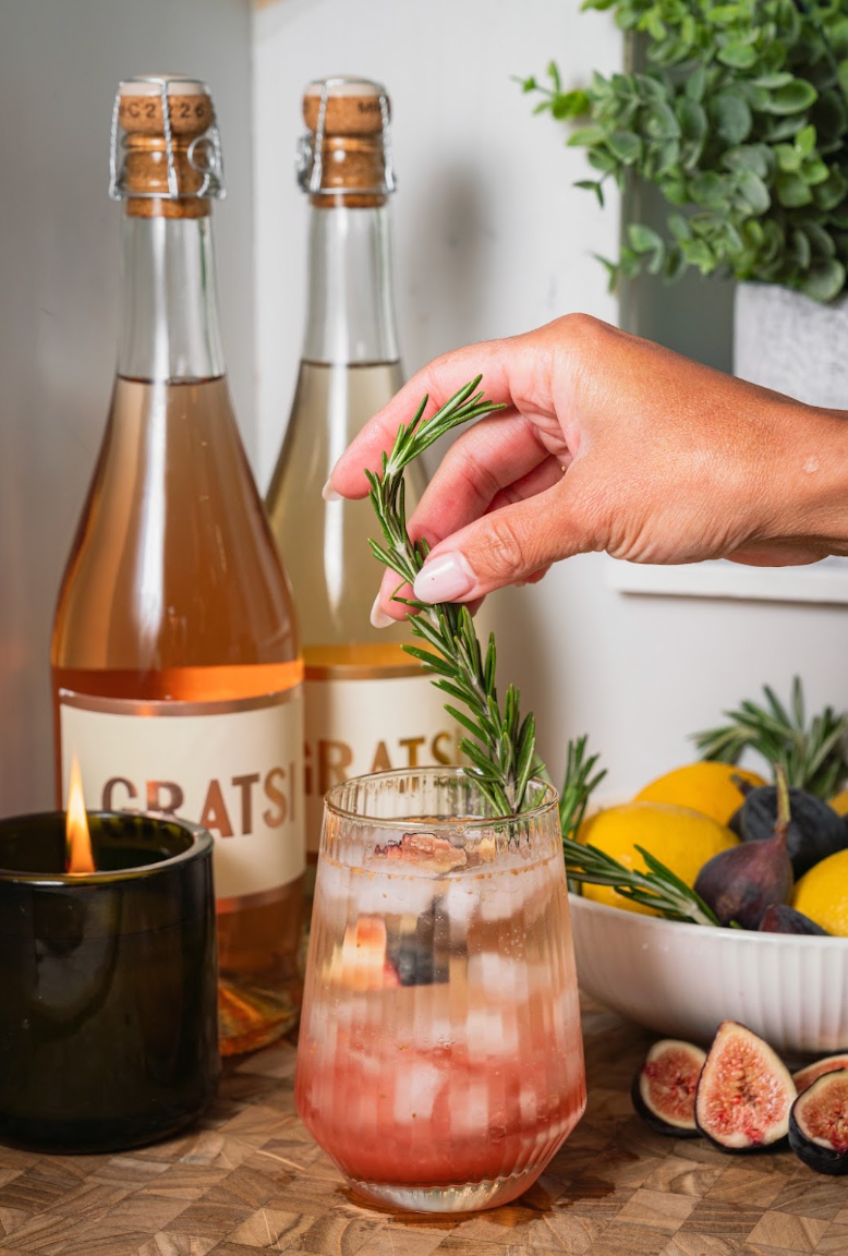 A hand places a sprig of rosemary into a glass of pink cocktail filled with ice, set next to two wine bottles, a lit candle, and a bowl of fresh figs and lemons.