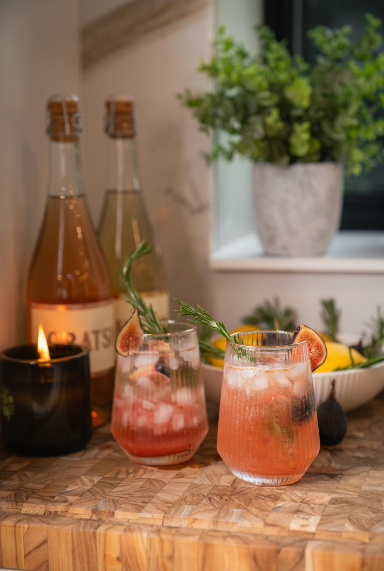 Two glasses of pink cocktails garnished with rosemary sprigs and fig slices, sitting on a wooden surface near bottles and a lit candle.