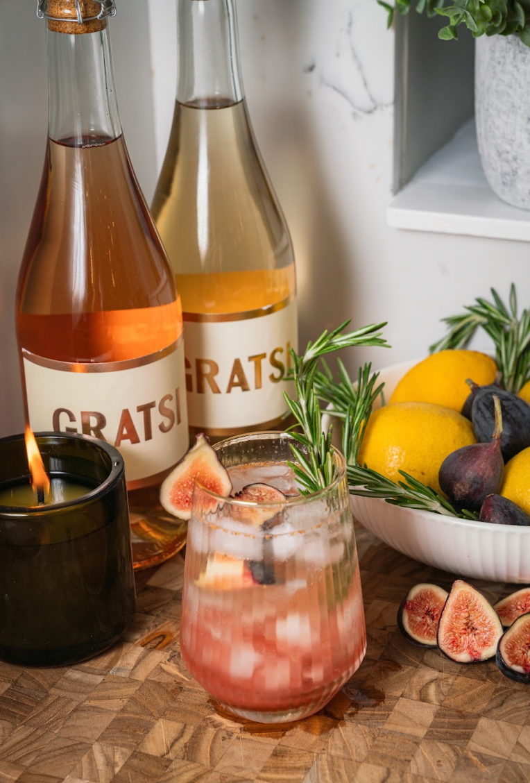A glass of pink cocktail garnished with a fig slice and rosemary, beside two Gratsi wine bottles, a bowl of lemons and figs, and a lit candle.