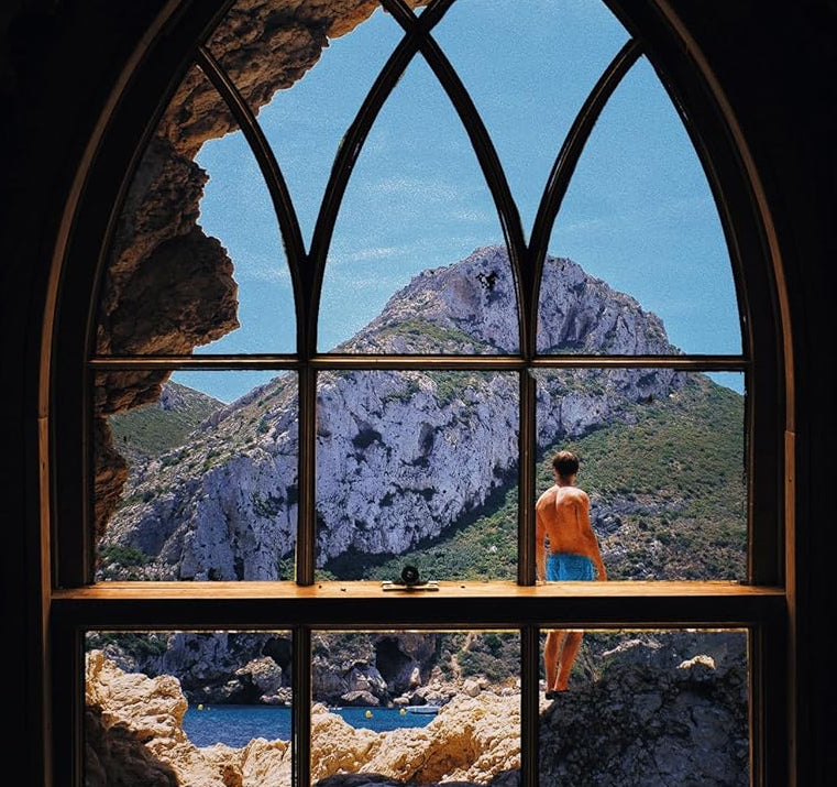 A man in blue shorts stands on rocky terrain outside a large arched window, looking at mountains under a clear blue sky, with blue water visible below.
