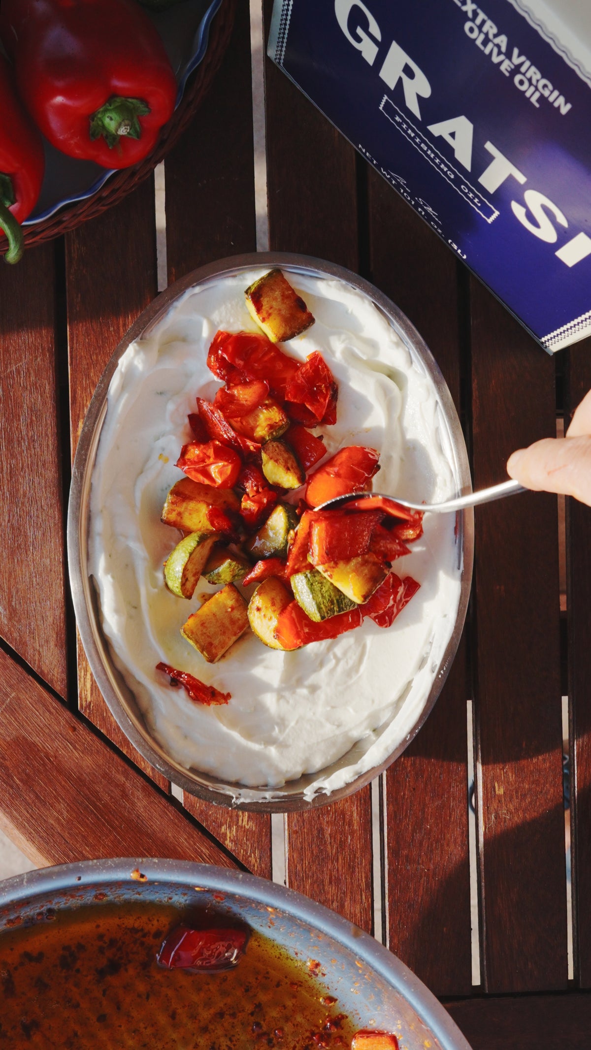 A hand uses a spoon to stir roasted red peppers and zucchini into a bowl of creamy dip.