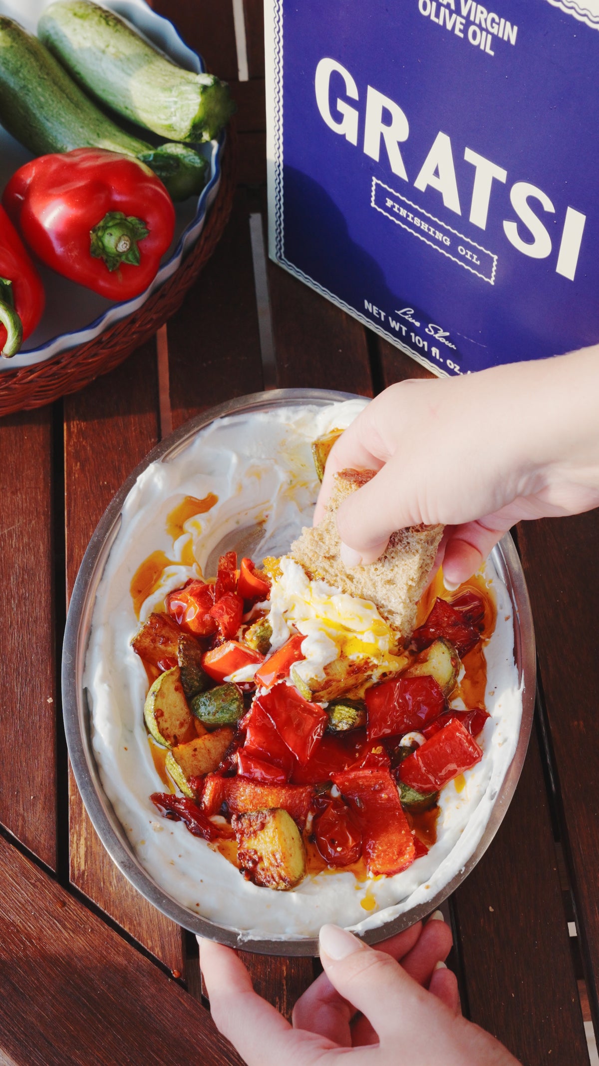 A hand dips a piece of bread into a bowl of yogurt topped with roasted red peppers and zucchini, next to a tin of Gratsi olive oil.