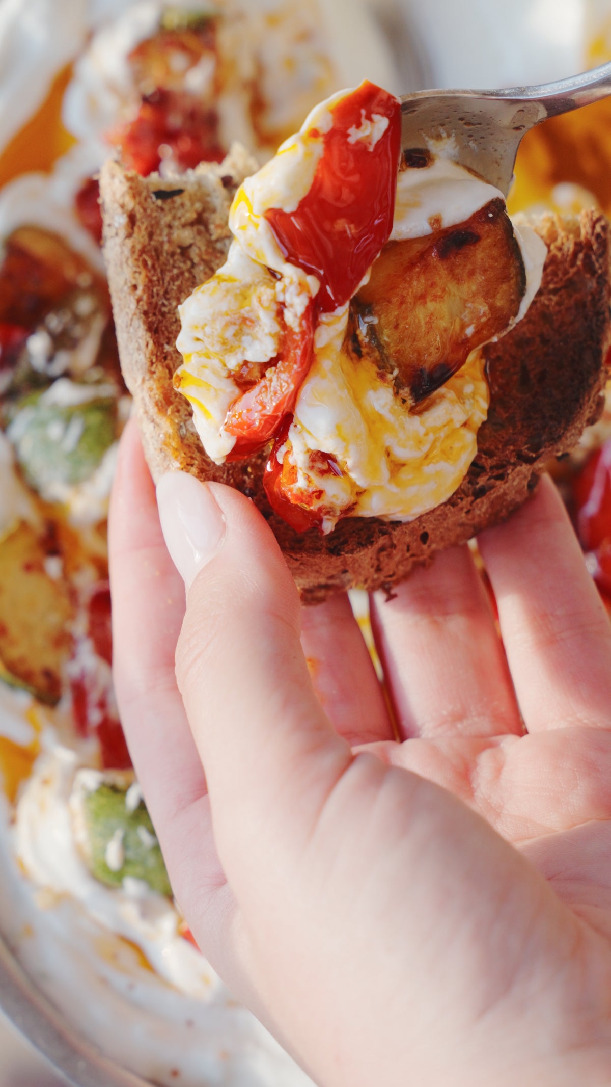 A hand holding a slice of toasted bread topped with roasted vegetables and creamy cheese.
