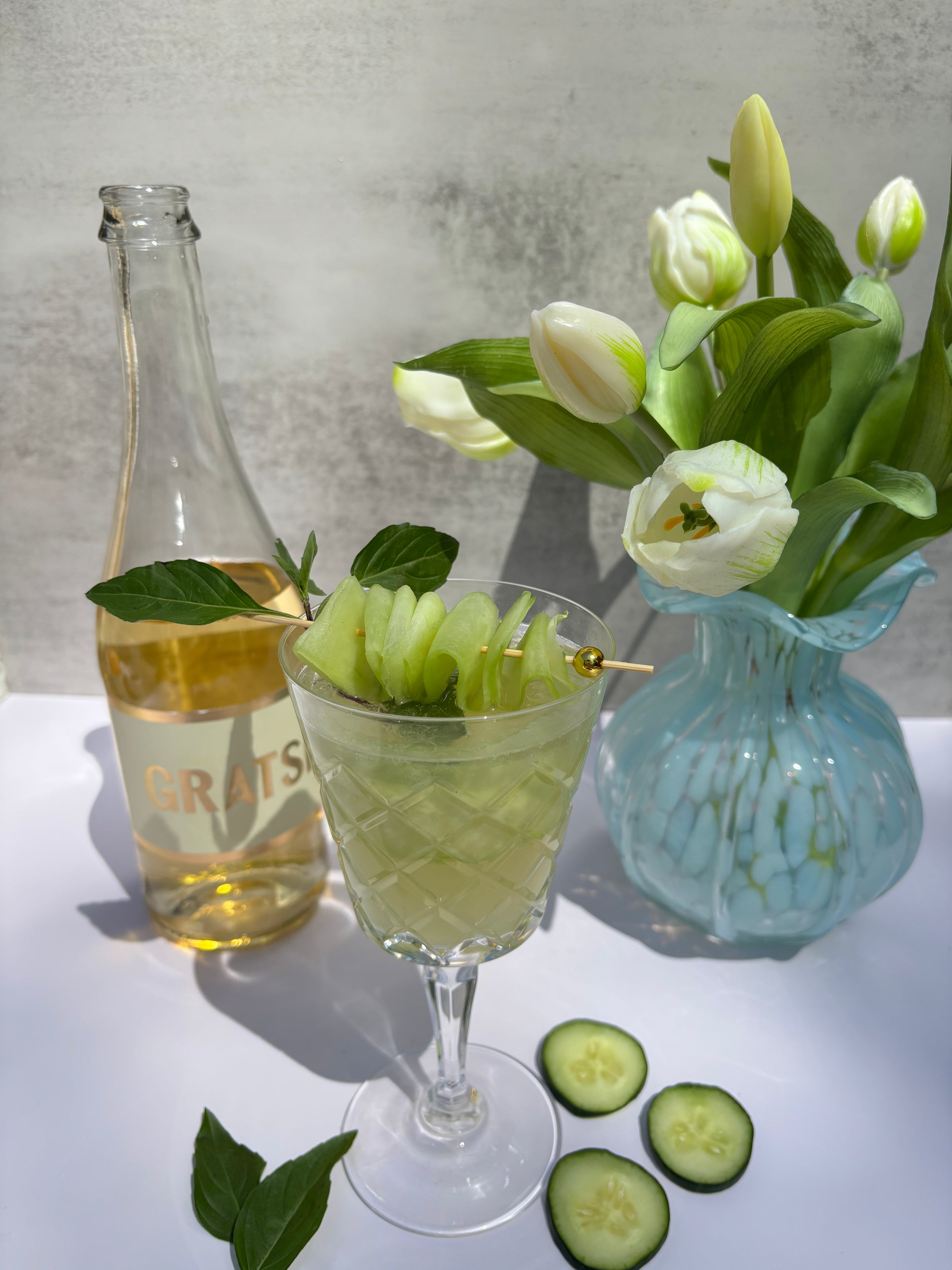 A glass of a light green cocktail garnished with cucumber slices and basil sits beside a bottle of GRATS wine and a blue vase with white tulips. Sliced cucumbers and basil leaves are on the white surface nearby.