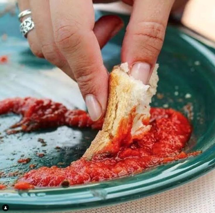 A hand dipping a piece of bread into a pool of red tomato sauce on a green plate, with only a few remnants of sauce left.