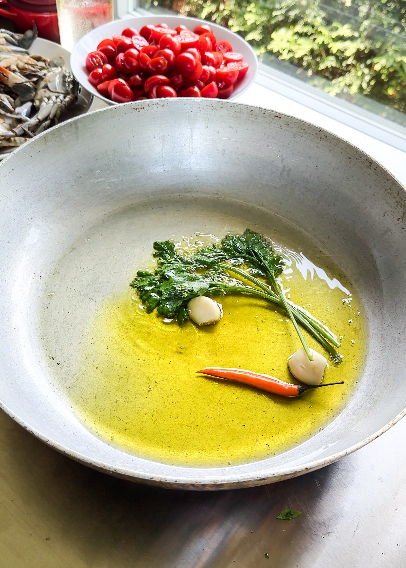 A pan with olive oil, garlic cloves, a red chili, and parsley on a kitchen counter. Nearby are a bowl of halved cherry tomatoes and a plate of raw shrimp. Sunlight streams through a window in the background.