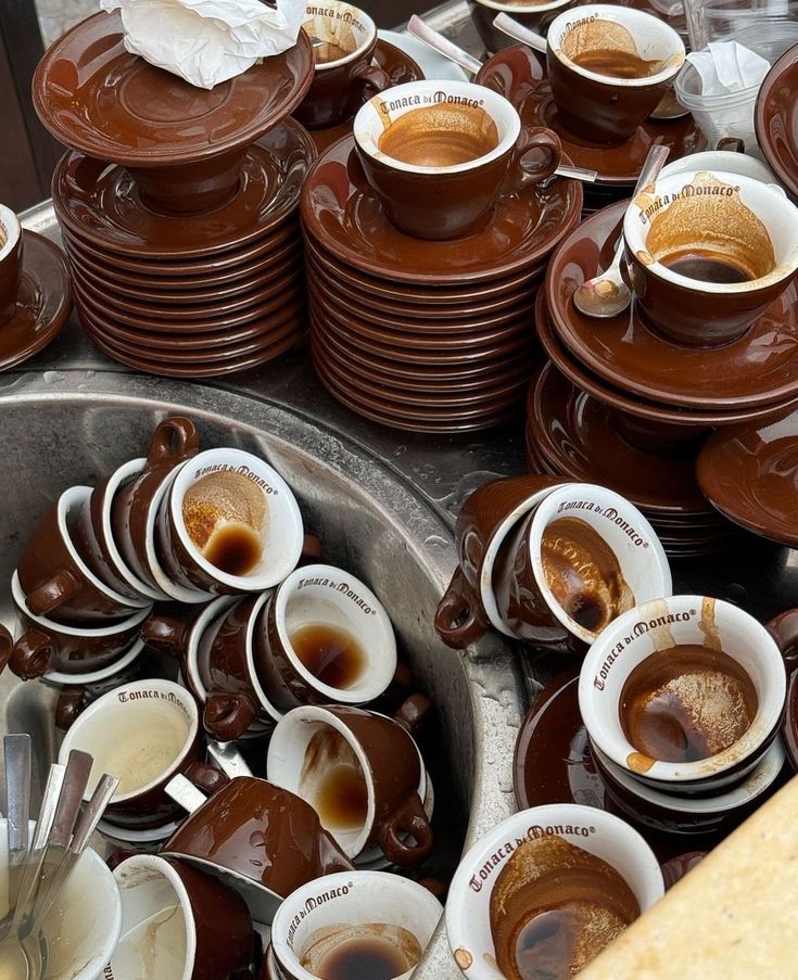 Stacks of brown espresso cups and saucers, many stained with leftover coffee, are piled around and inside a sink. Some cups have spoons, and a paper napkin sits atop one pile.