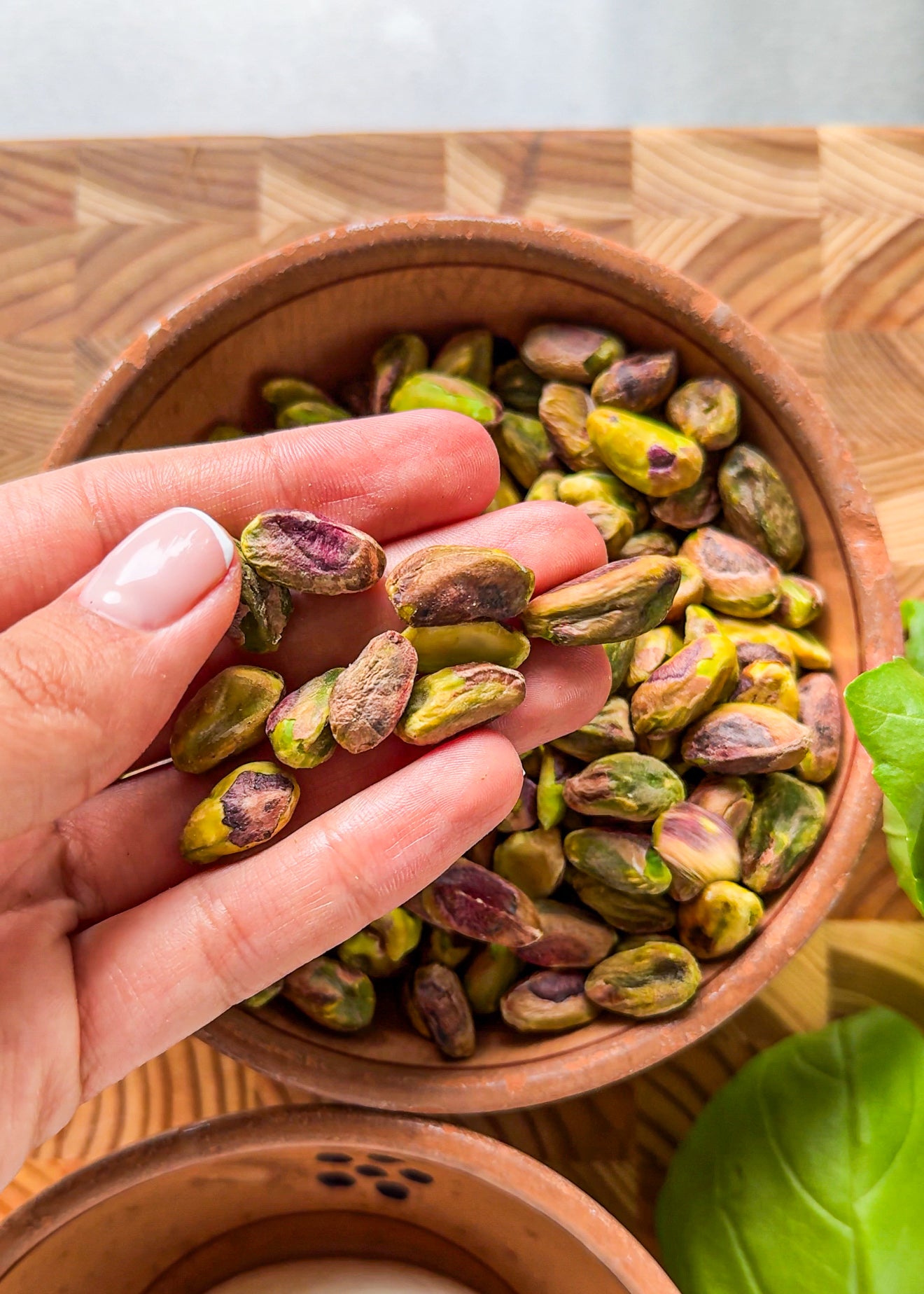 A hand holds several shelled pistachios above a wooden bowl filled with more pistachios, with green leaves and another bowl nearby on a wooden surface.