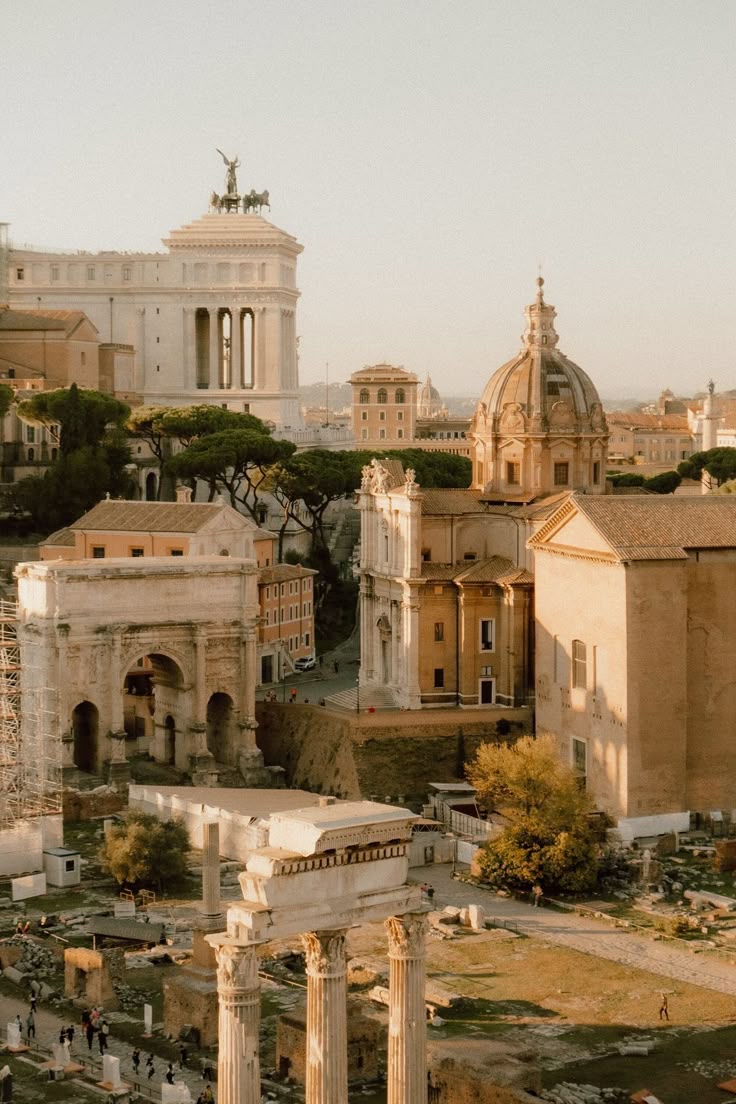 A warm, sunlit view of ancient Roman ruins and historic buildings in Rome, Italy, featuring columns, arches, domed roofs, and the Victor Emmanuel II Monument in the background. People walk among the ruins.
