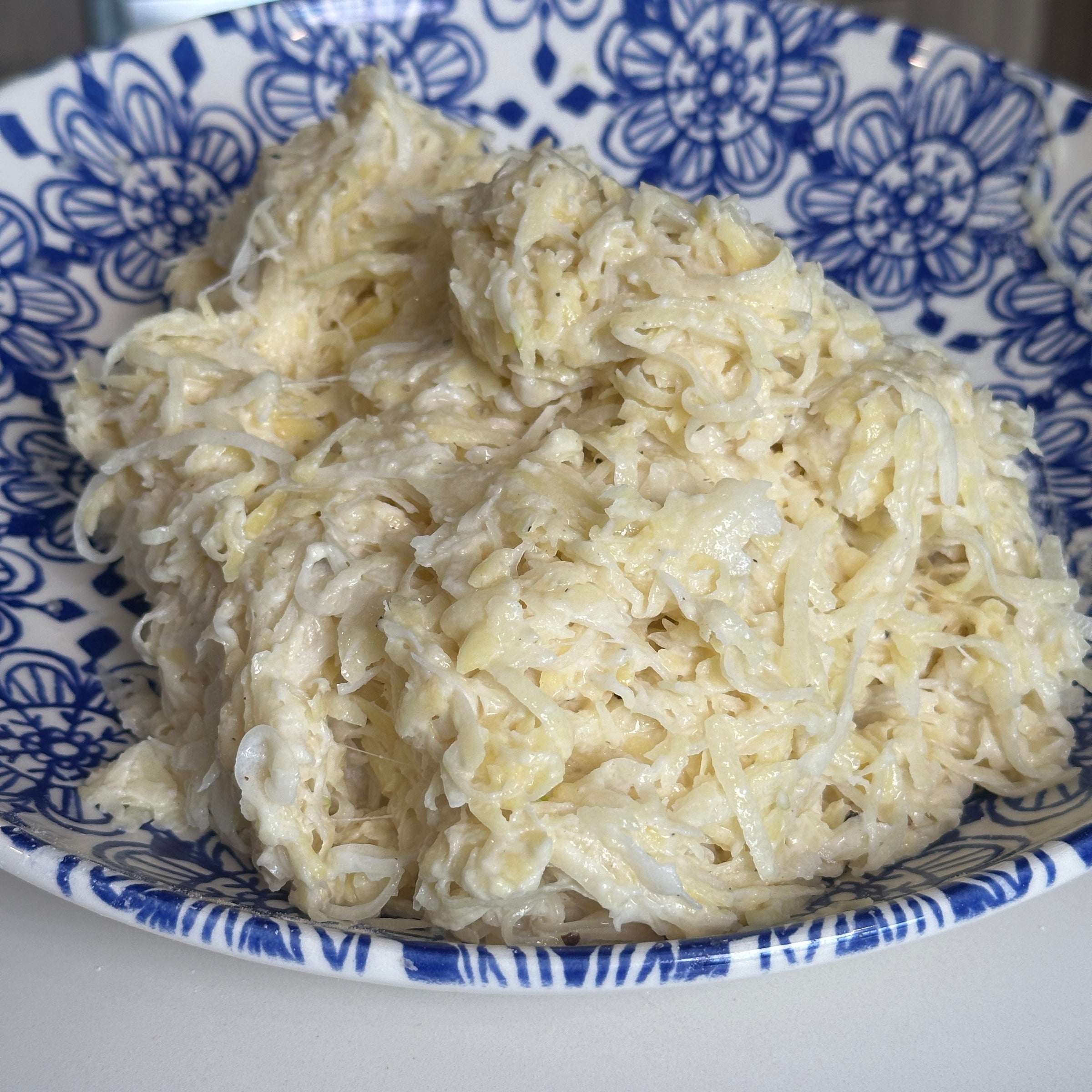 A blue and white patterned bowl filled with a creamy shredded cabbage salad, sitting on a white surface.