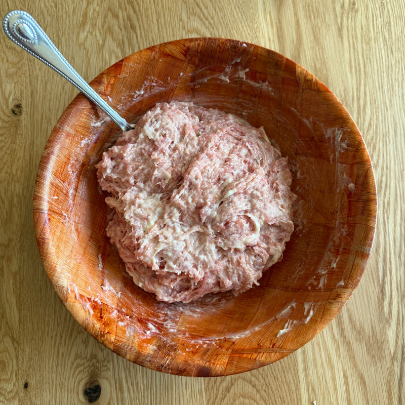 A wooden bowl on a light wood table contains a mixture of ground meat and other ingredients with a metal spoon resting inside the bowl.