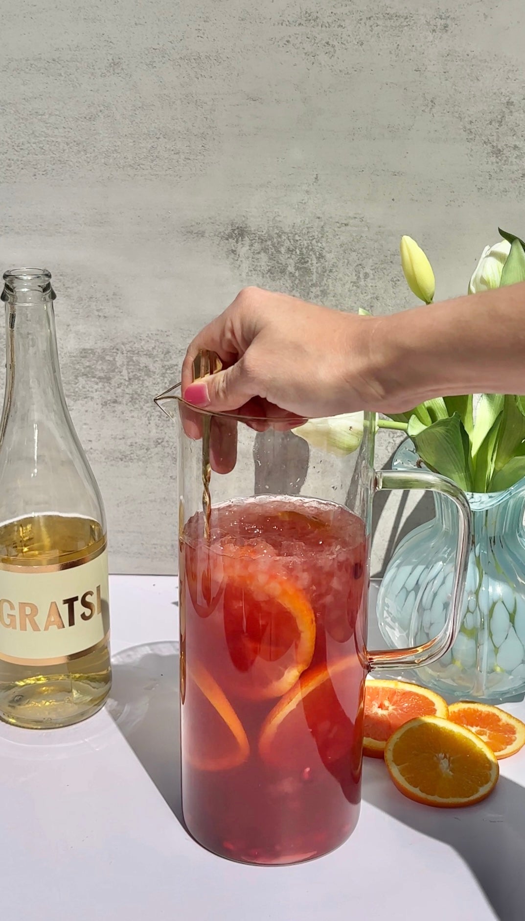A hand stirs a large glass pitcher filled with iced red drink and orange slices. Beside it are a bottle labeled GRATS, a glass vase with white flowers, and sliced oranges on a white surface.