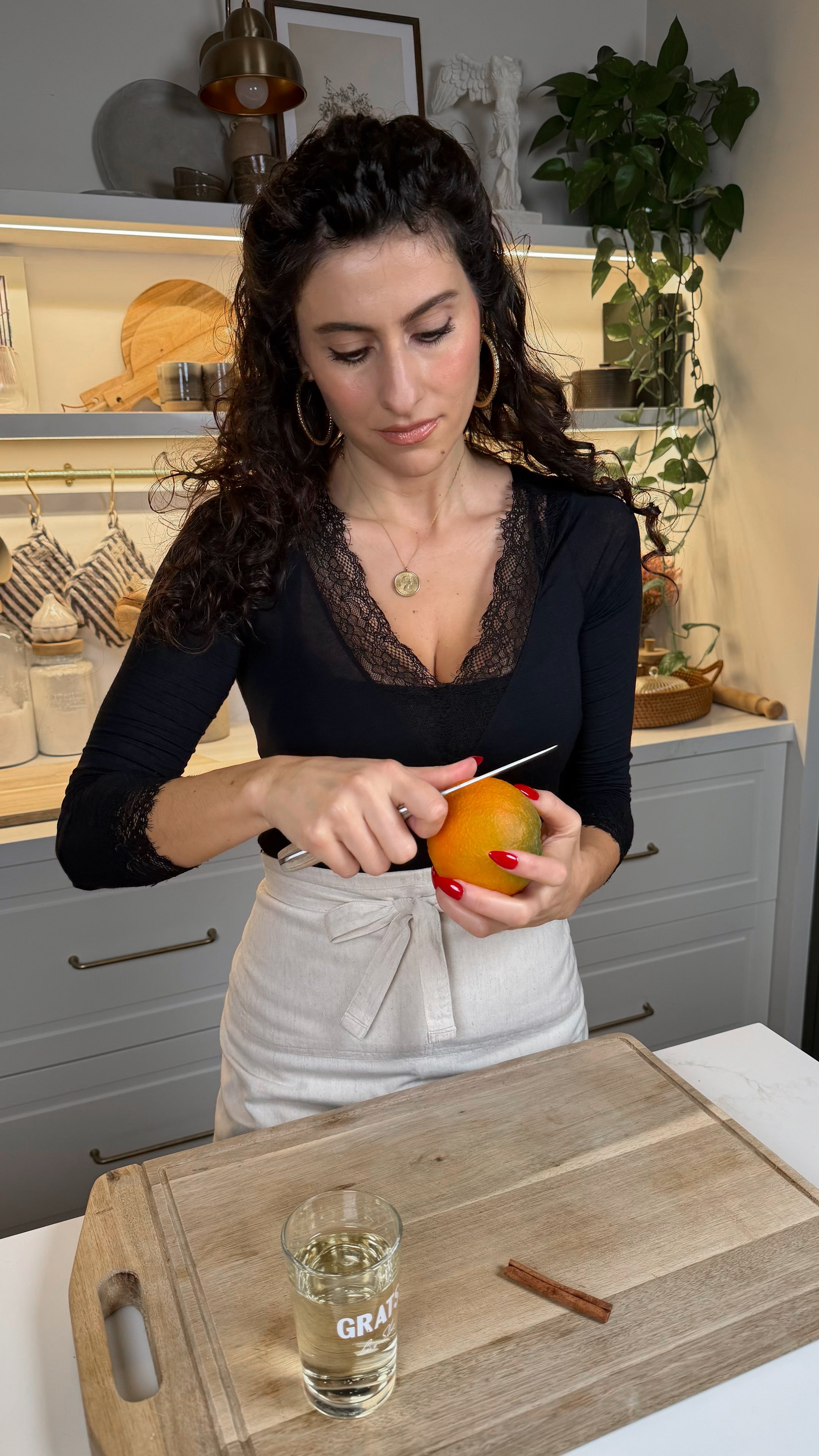 A woman with curly dark hair, wearing a black top and white pants, stands in a kitchen slicing an orange with a knife over a wooden cutting board. A glass and a cinnamon stick are on the board.