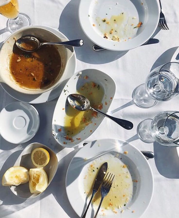Overhead view of a white tablecloth with empty plates, dirty utensils, used glasses, lemon wedges, and leftover food remnants, suggesting a recently finished meal.