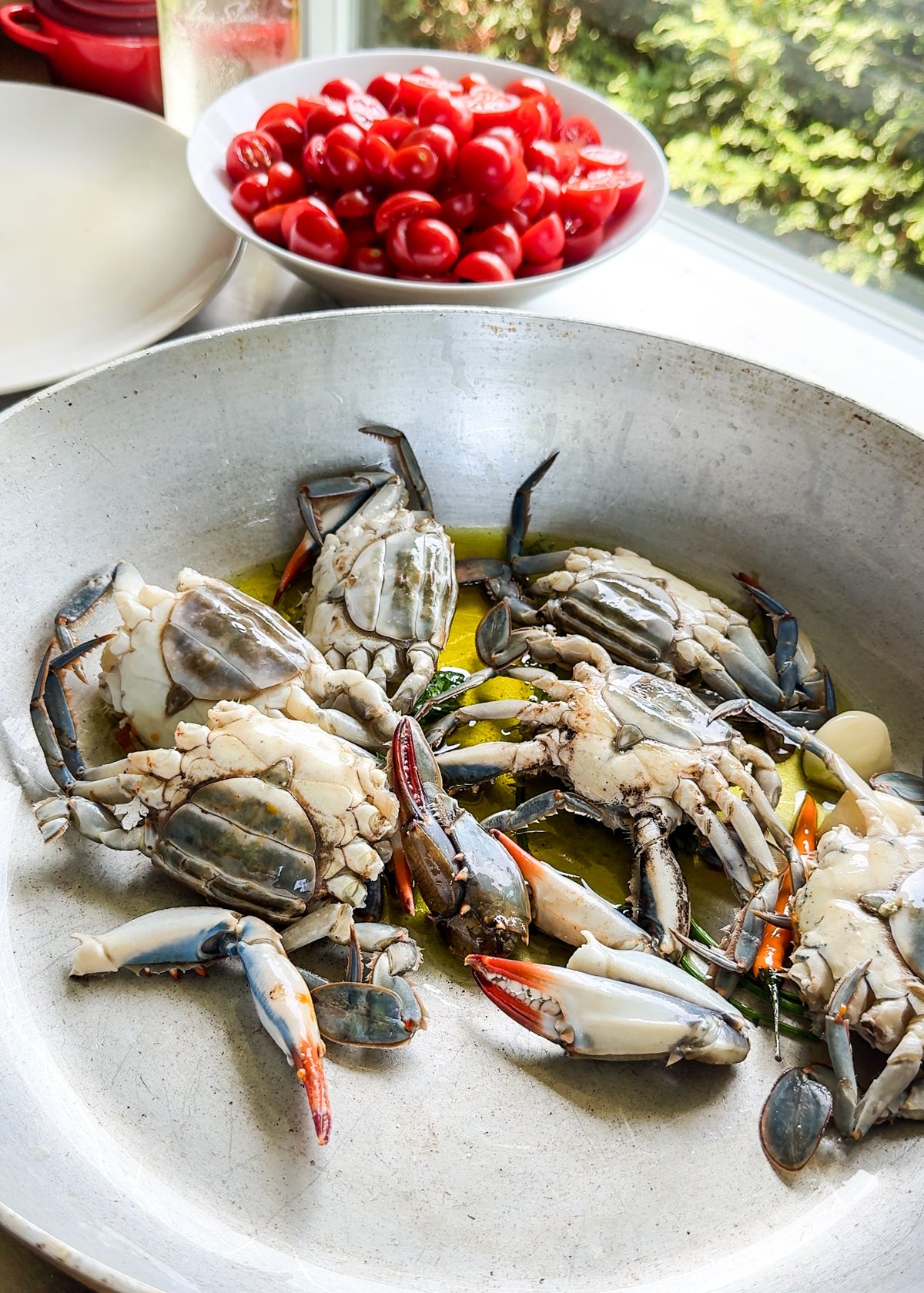 Raw crabs in a metal pan with oil, garlic, and herbs. In the background, a bowl of red grape tomatoes sits on a windowsill with green trees visible outside.
