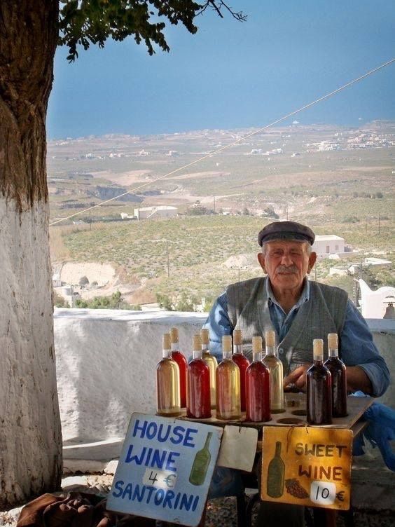 An older man sits at a small outdoor table displaying bottles of red and white wine for sale, with handwritten signs reading House Wine Santorini 4€ and Sweet Wine 10€. A rural landscape is in the background.