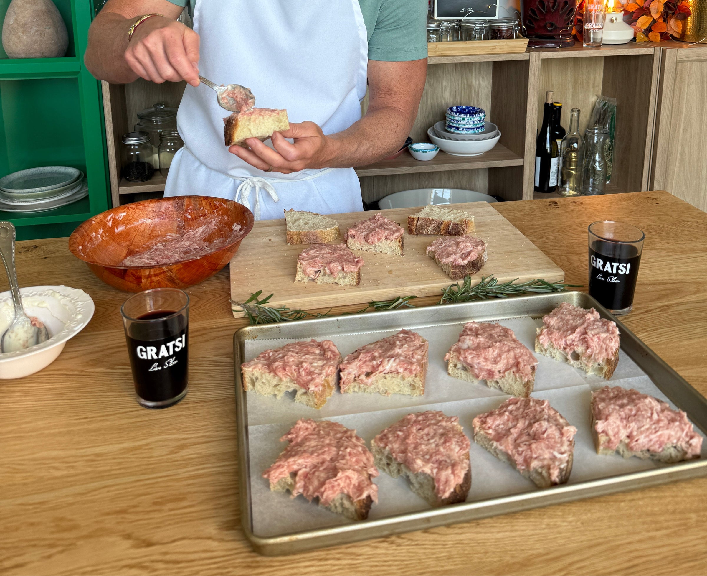 A person in an apron spreads a pink meat mixture onto slices of bread at a kitchen counter. Several slices are prepared on a tray and wooden board. Two glasses labeled GRATSI are on the table.