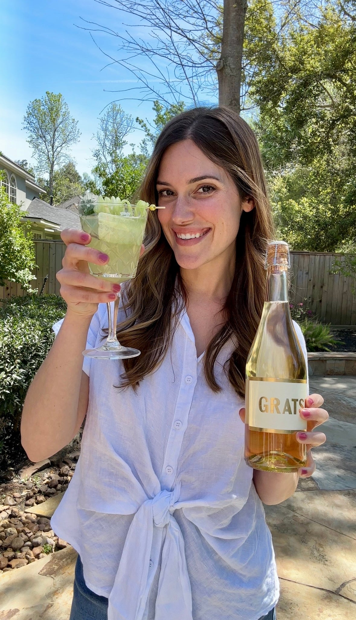 A woman standing outside smiles while holding a glass of a green beverage in one hand and a bottle labeled GRATS in the other. She is wearing a white shirt and is in a sunny backyard with trees and a fence.