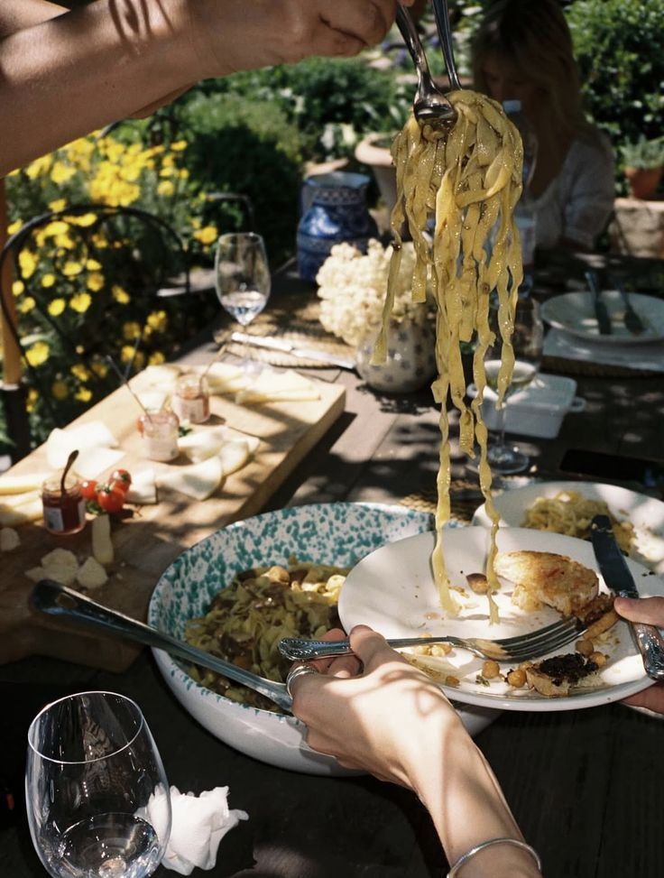 A hand serves fresh pasta onto a plate at an outdoor table set with wine glasses, cheese, and appetizers, surrounded by greenery and sunlight.