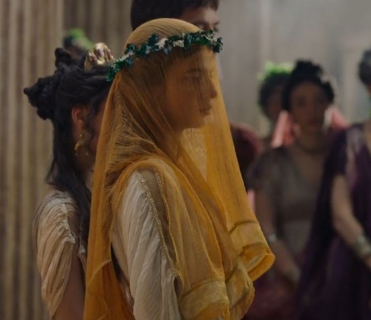 A woman wearing a yellow veil and a green leafy crown stands among others in ancient-style clothing, with soft lighting and blurred figures in the background.