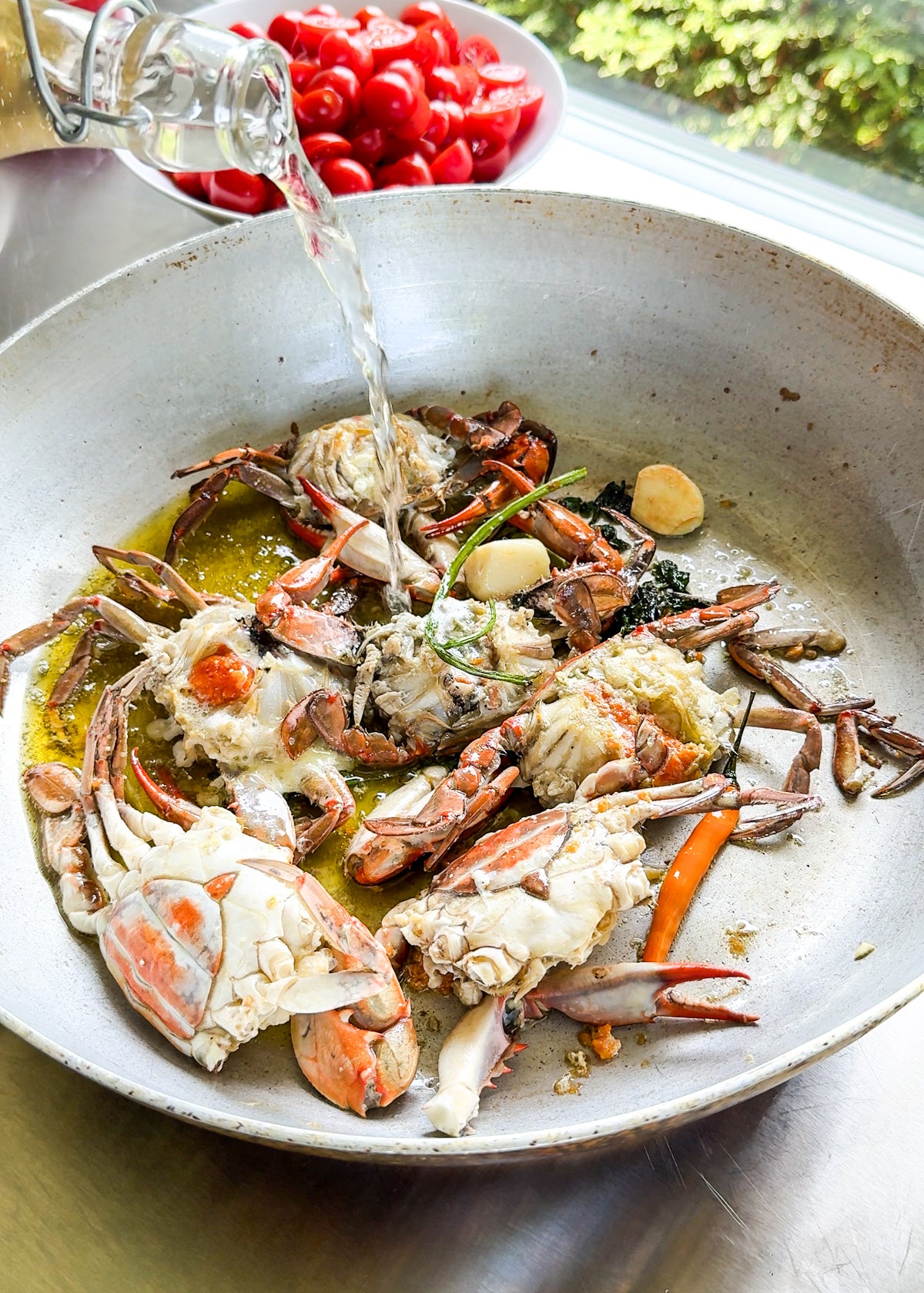 Raw blue crabs, garlic, herbs, and oil cook in a pan as water is poured in. A bowl of halved cherry tomatoes sits nearby on the counter next to a sunny window with green foliage outside.