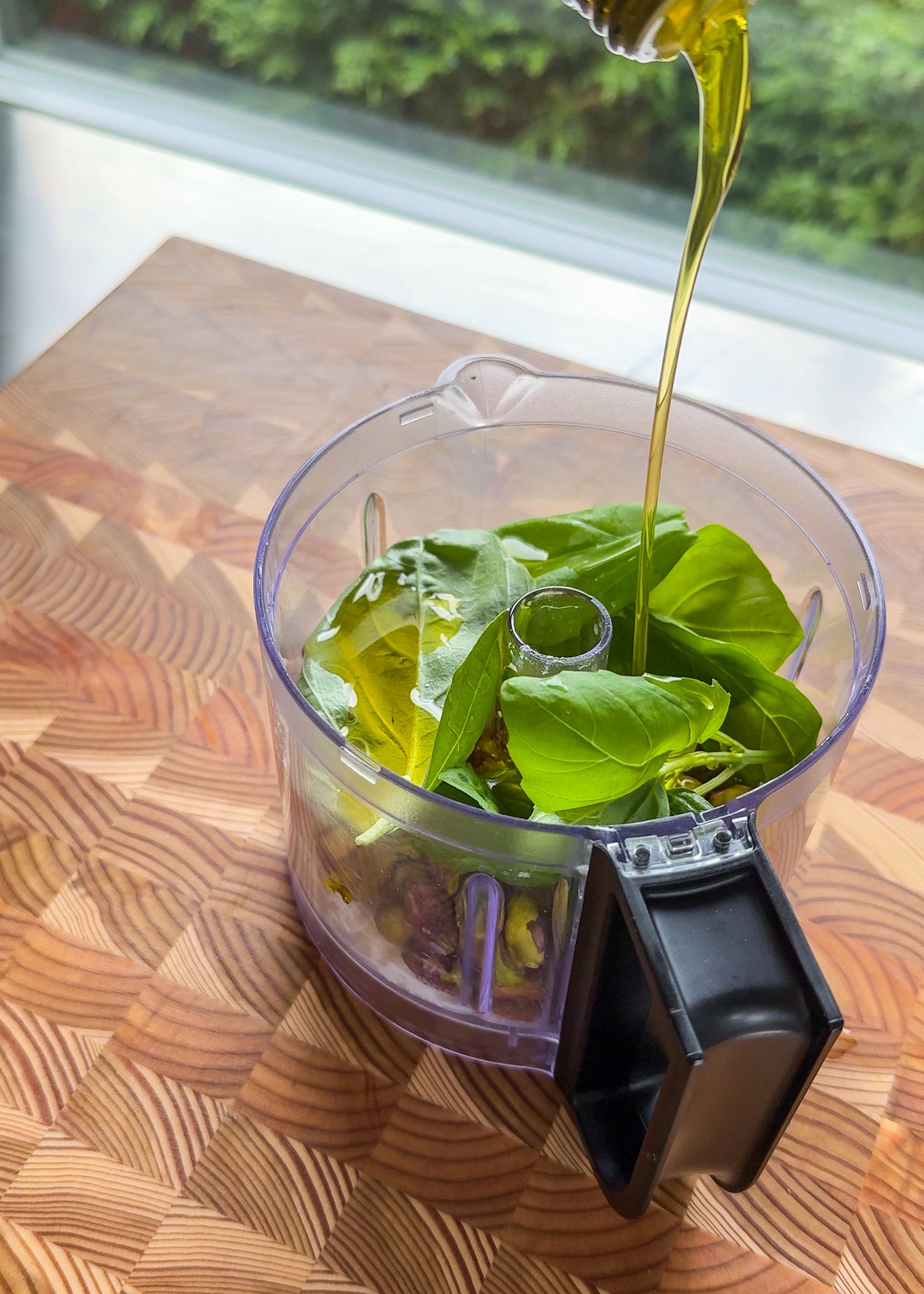 Olive oil is being poured into a food processor bowl with fresh green basil leaves, nuts, and garlic, all placed on a wooden surface near a window.
