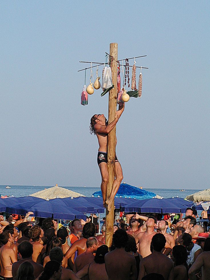 A man climbs a tall wooden pole on a crowded beach, reaching for prizes like food and bags hanging at the top, while people watch from below. Blue umbrellas and the sea are visible in the background.