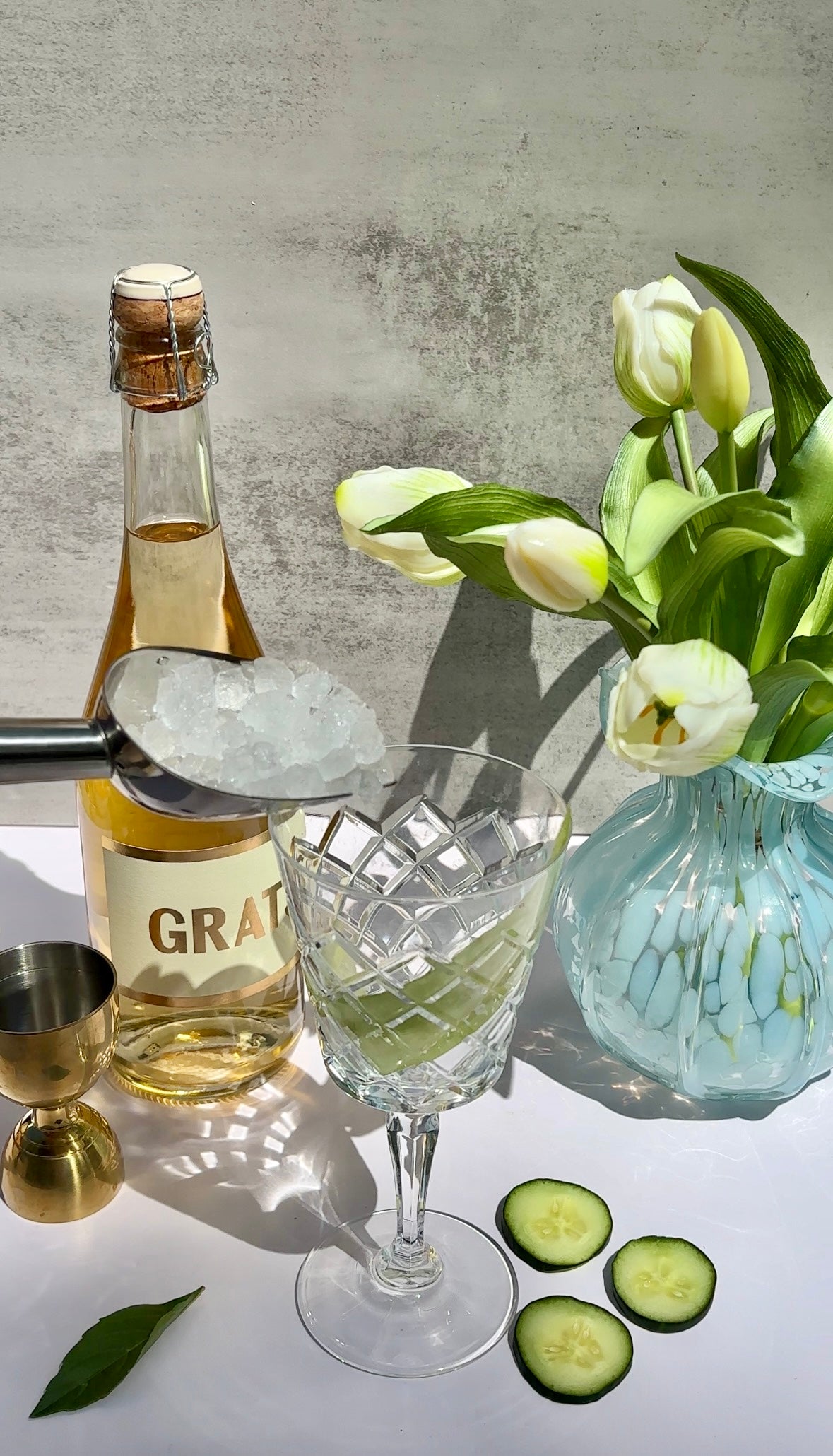 A crystal glass with a light green drink is being topped with ice using tongs. Nearby are a bottle labeled GRATA, a gold jigger, cucumber slices, a green leaf, and a vase of white tulips.