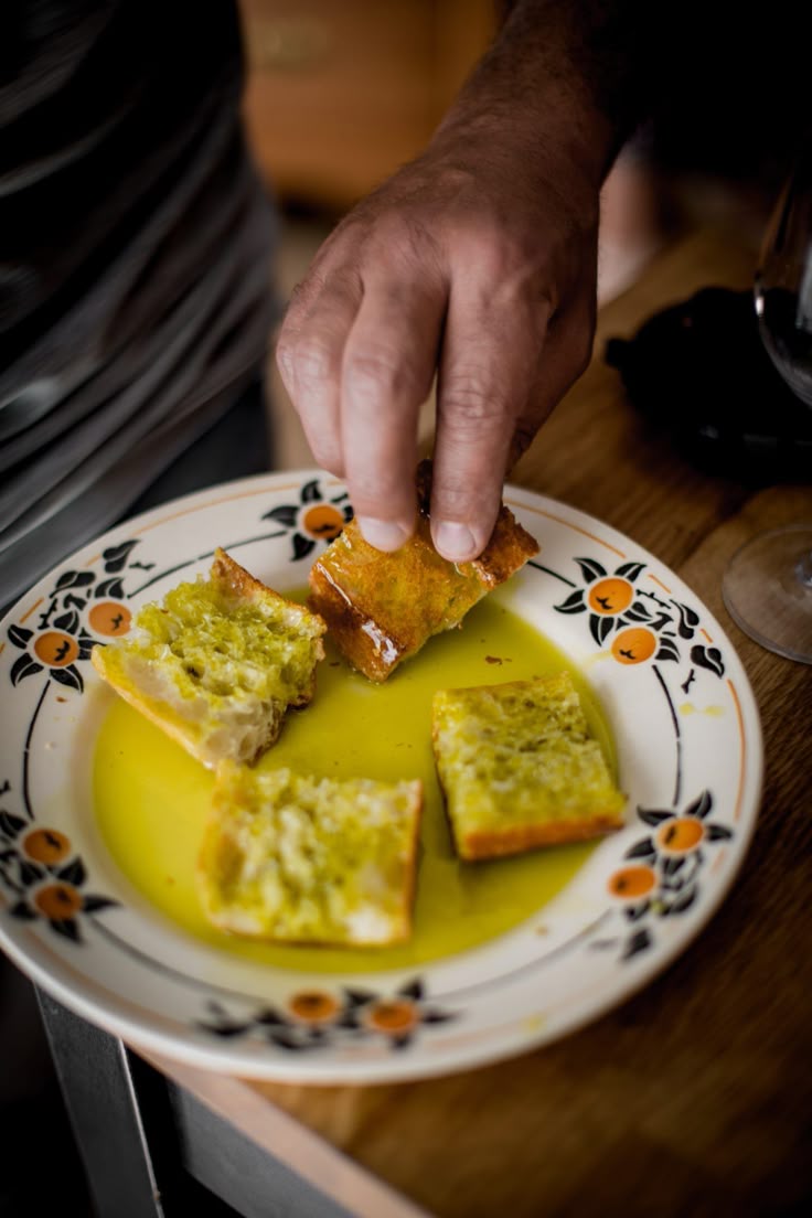 A hand dips a piece of bread into olive oil on a decorative plate with four pieces of bread soaking in the oil.