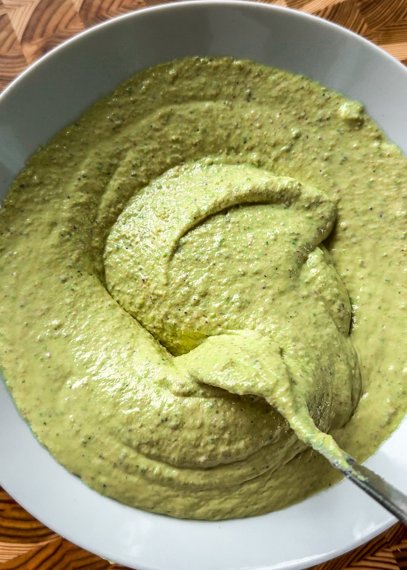 A close-up of a bowl filled with creamy green sauce or dip, with a spoon partially submerged, showing a thick, smooth texture. The background shows a wooden surface.