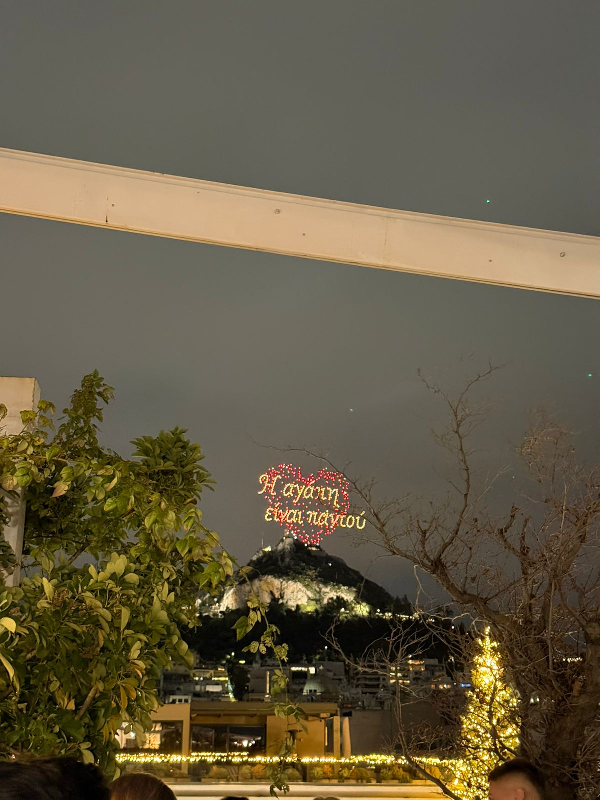 A nighttime scene with a lit Greek phrase in the sky, framed by trees, festive string lights, and a distant hilltop building. The sky is cloudy, and the scene feels warm and festive.