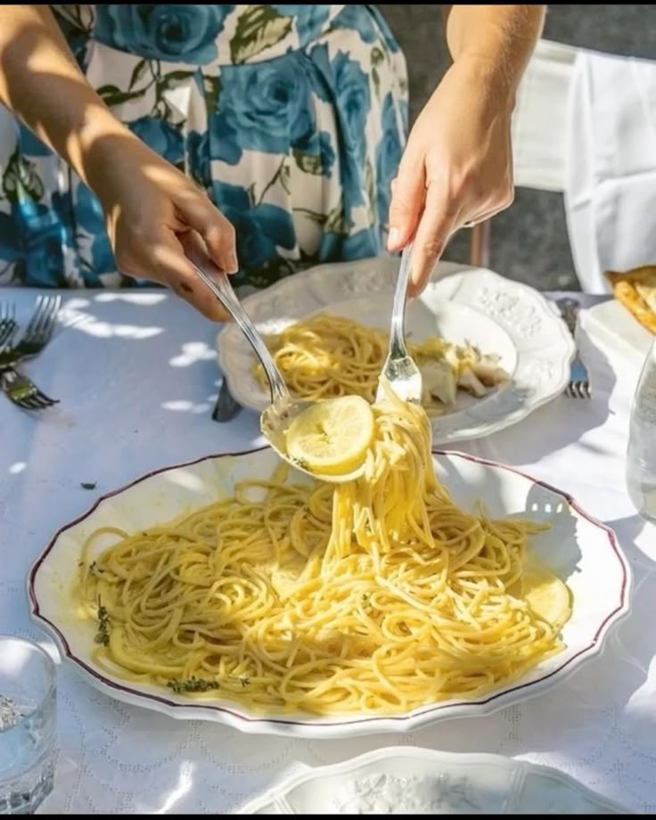 A person in a blue floral dress is serving lemon pasta from a large white dish onto a plate at an outdoor table set for a meal. Sunlight brightens the scene.