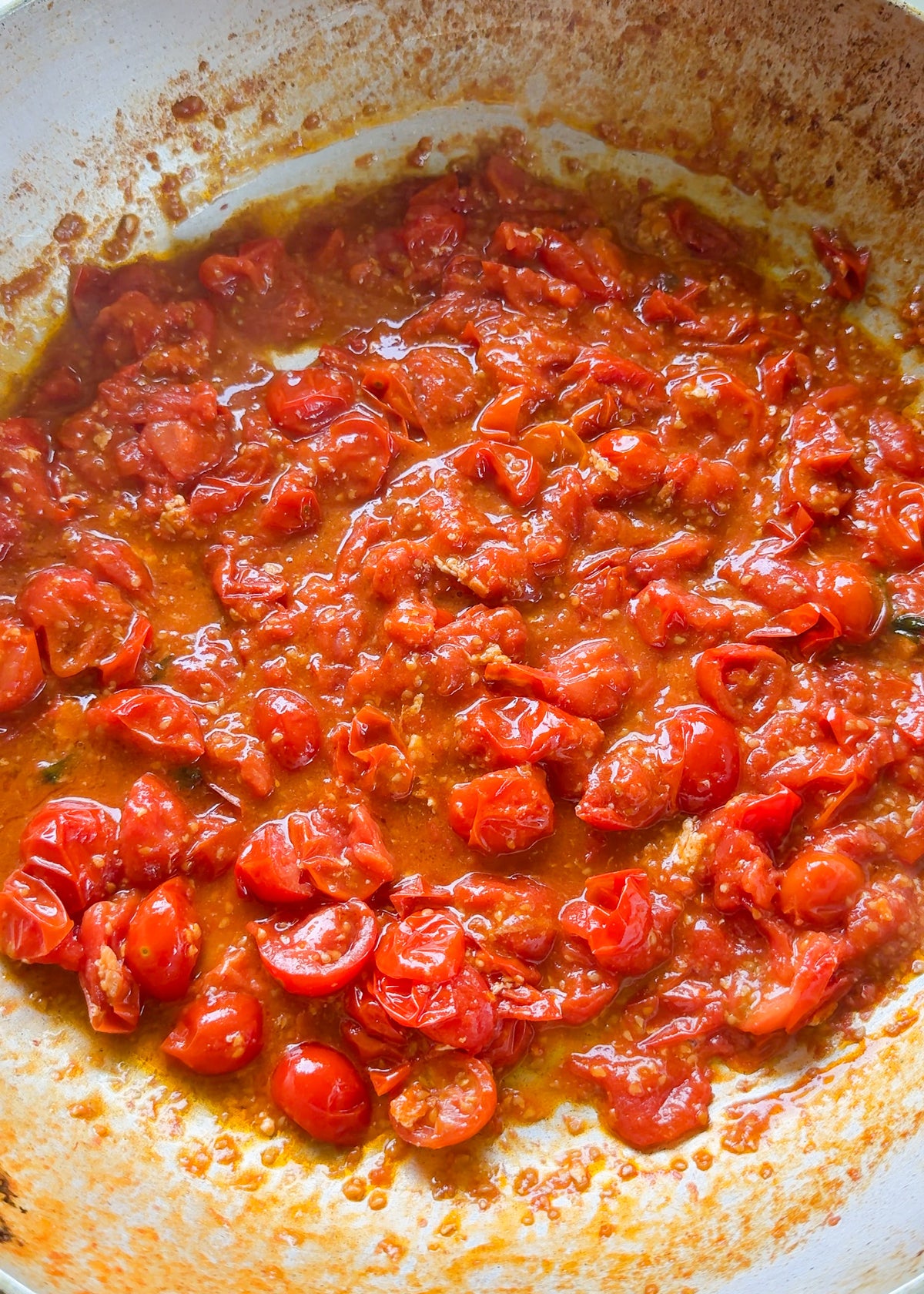Cherry tomatoes cooking in a skillet, breaking down into a chunky, red tomato sauce with visible seeds and juices, surrounded by oil and spices.