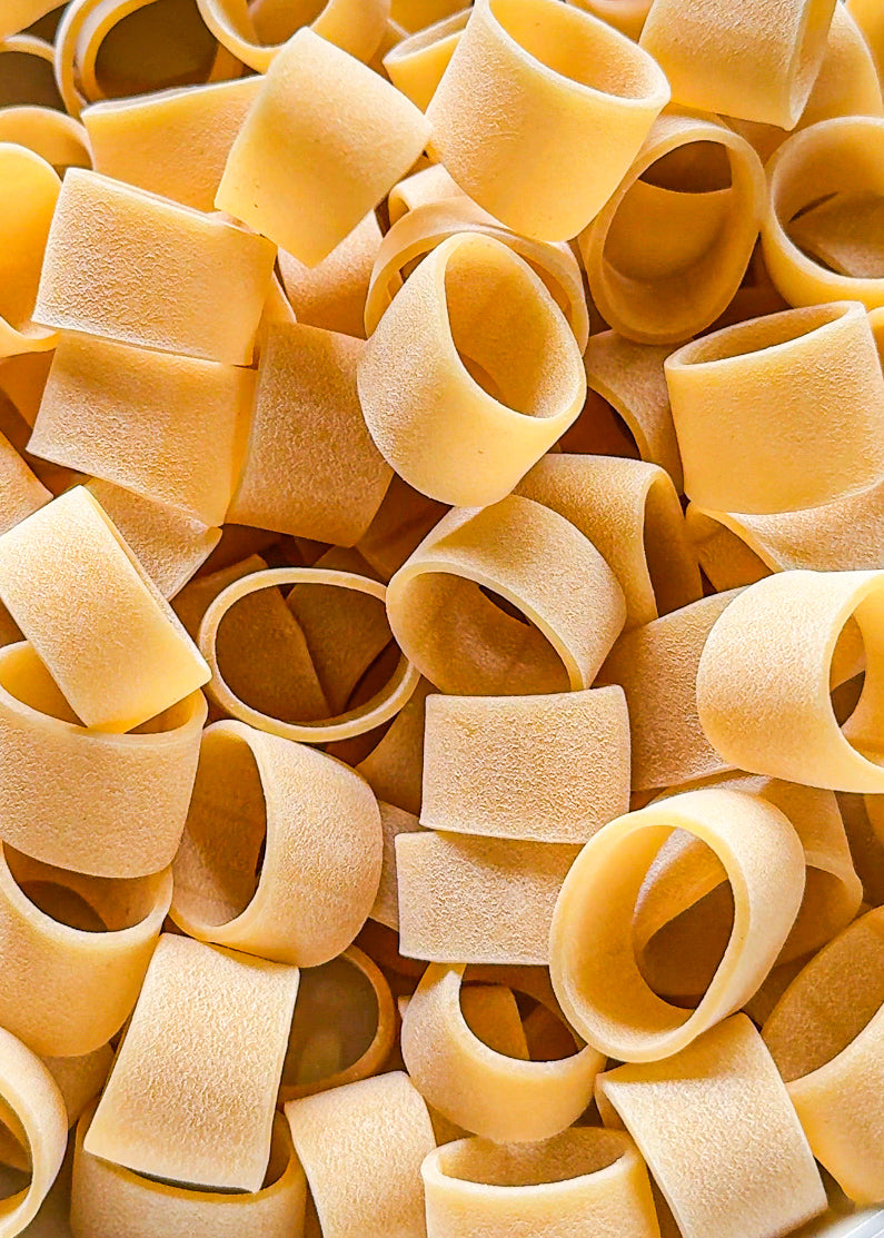 Close-up of uncooked short pasta tubes, possibly paccheri or mezzi paccheri, tightly packed together in a random arrangement. The surface texture is slightly rough and matte.