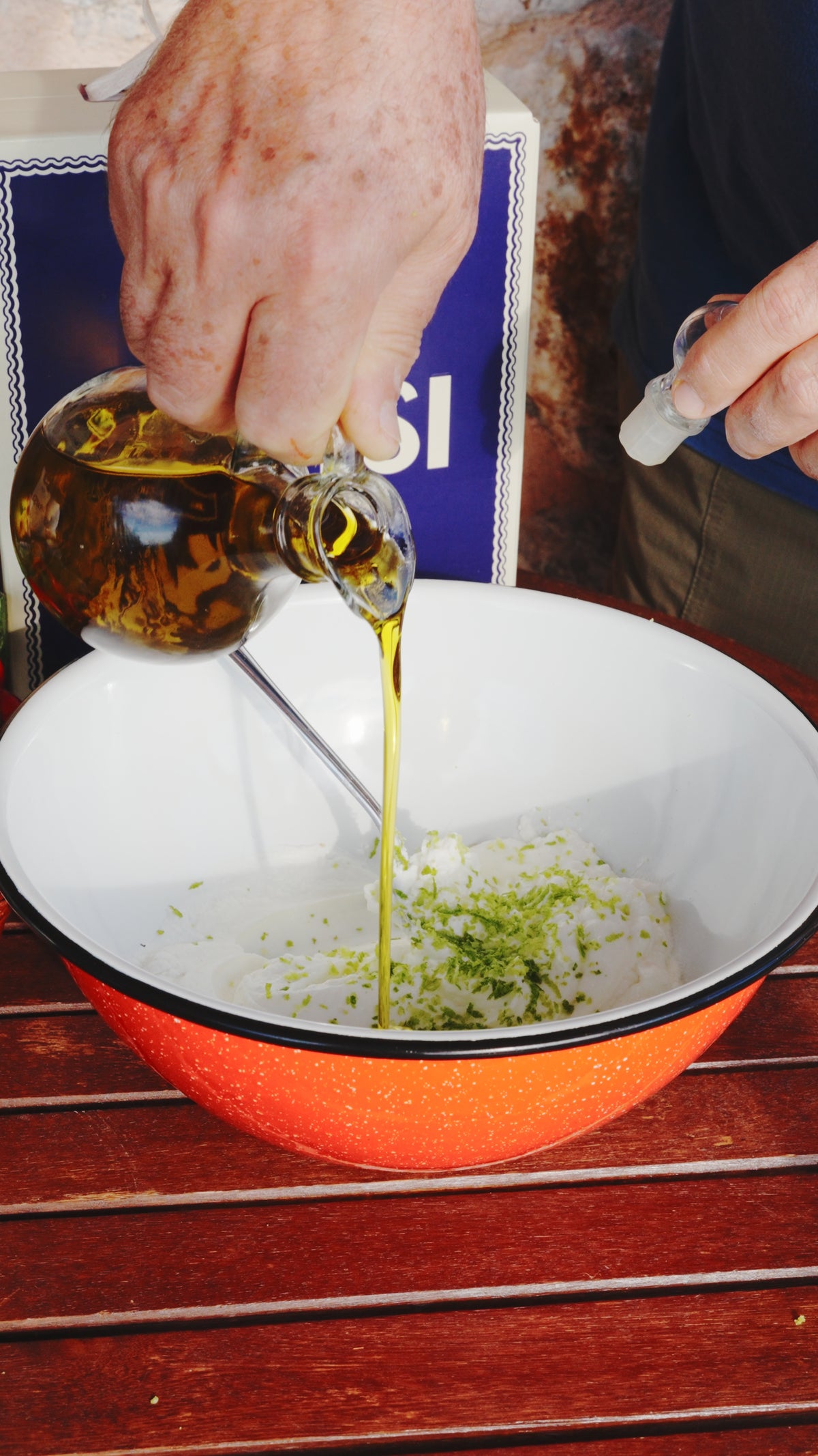 A person pours olive oil from a glass carafe into a mixing bowl containing white cream and lime zest.