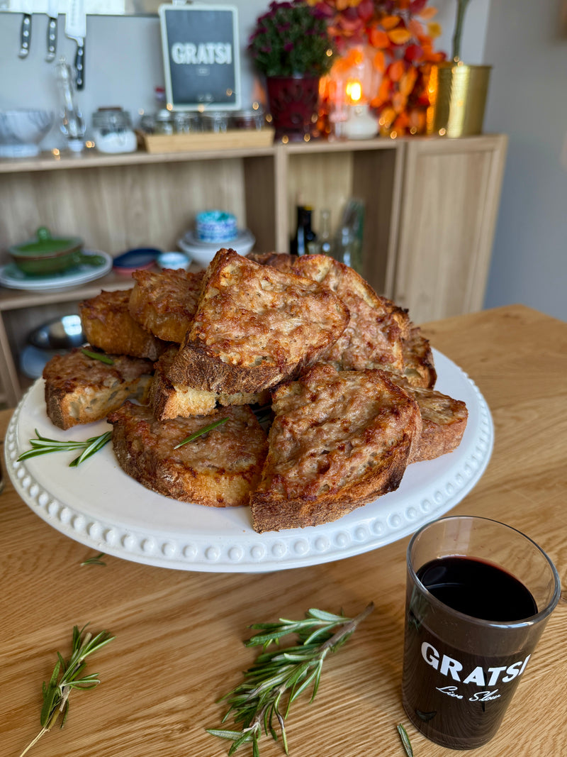 A plate of toasted bread slices with melted cheese sits on a wooden table, garnished with rosemary. A glass of red wine labeled GRATSI is in front, with shelves and autumn decor in the background.