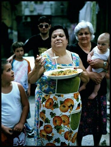 A woman in a colorful apron holds a plate of food, surrounded by children and an older woman holding a baby. The group stands together outdoors, appearing to enjoy a communal or family gathering.