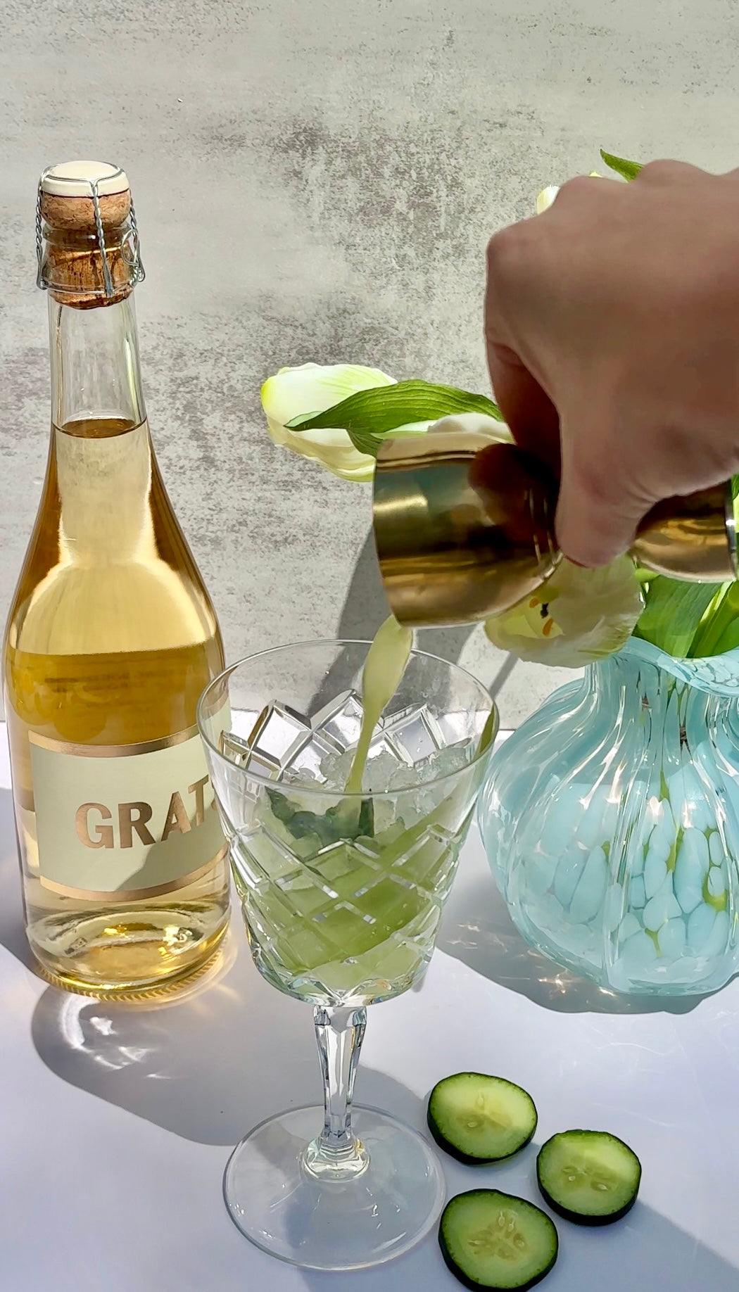A hand pours liquid from a gold jigger into a crystal glass with a light cocktail. Nearby are a bottle labeled “GRATI,” a blue vase with white flowers, and cucumber slices on a white surface.