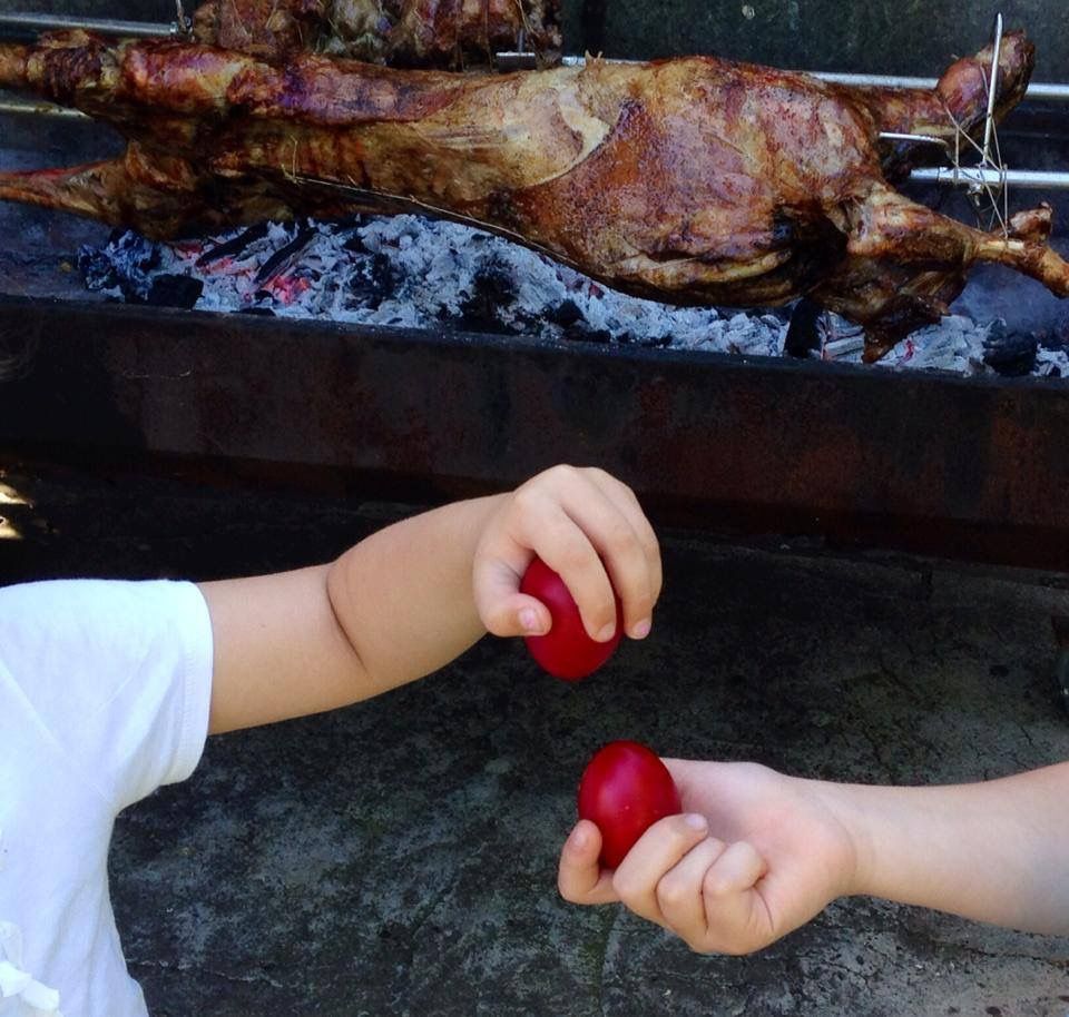 Two children holding red dyed eggs are about to tap them together, with a whole roasted lamb on a spit cooking over coals in the background.