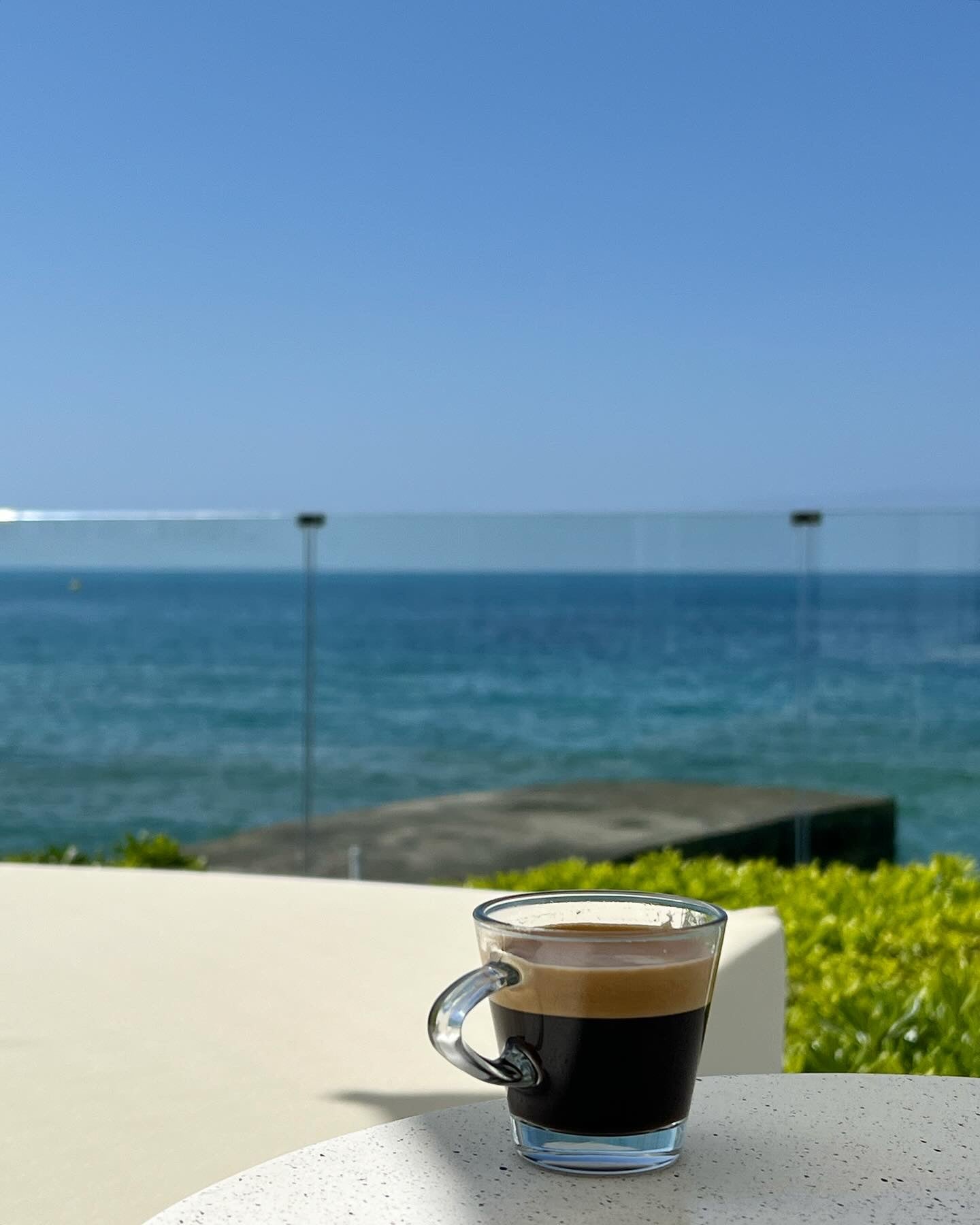 A clear glass cup of black coffee sits on a white table outdoors, overlooking a calm blue sea with a clear sky and a glass railing in the background.