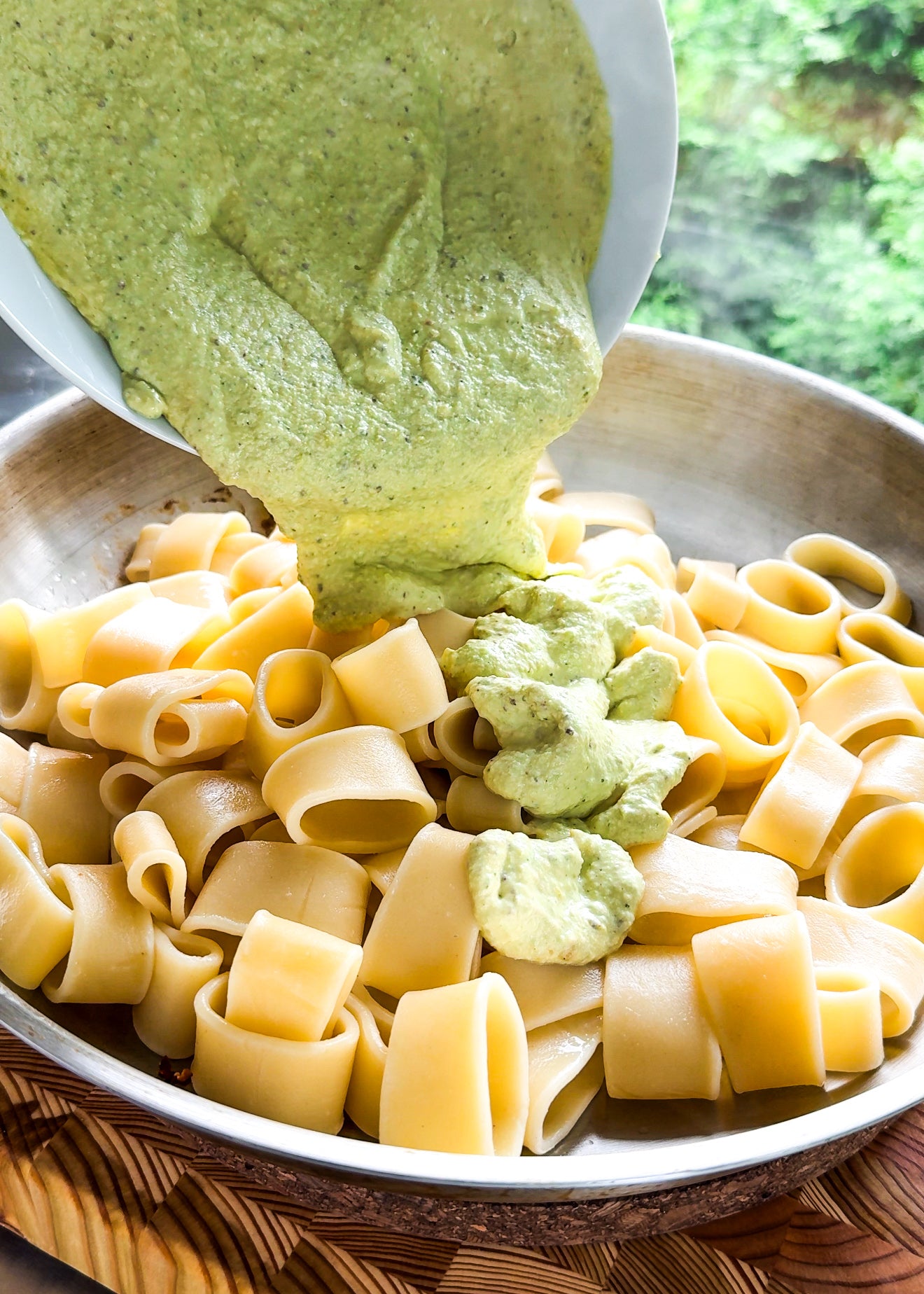 A creamy green sauce is being poured from a white bowl over cooked pasta in a pan, with a blurred green background visible behind the dish.