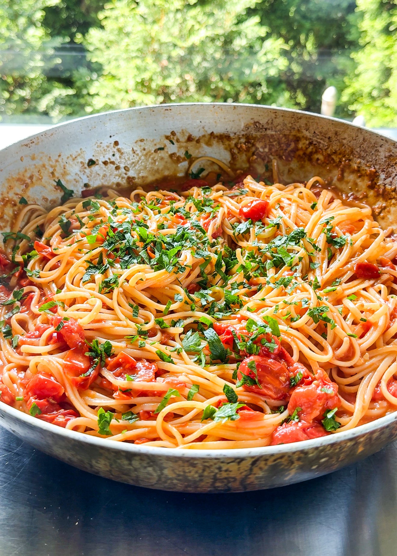 A skillet filled with spaghetti topped with chopped fresh herbs and tomato sauce sits on a table, with greenery visible in the bright background.